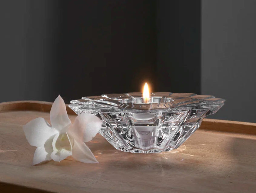 A clear glass tealight holder with a lit flame, placed on a wooden tray next to a white orchid flower against a dark gray background.