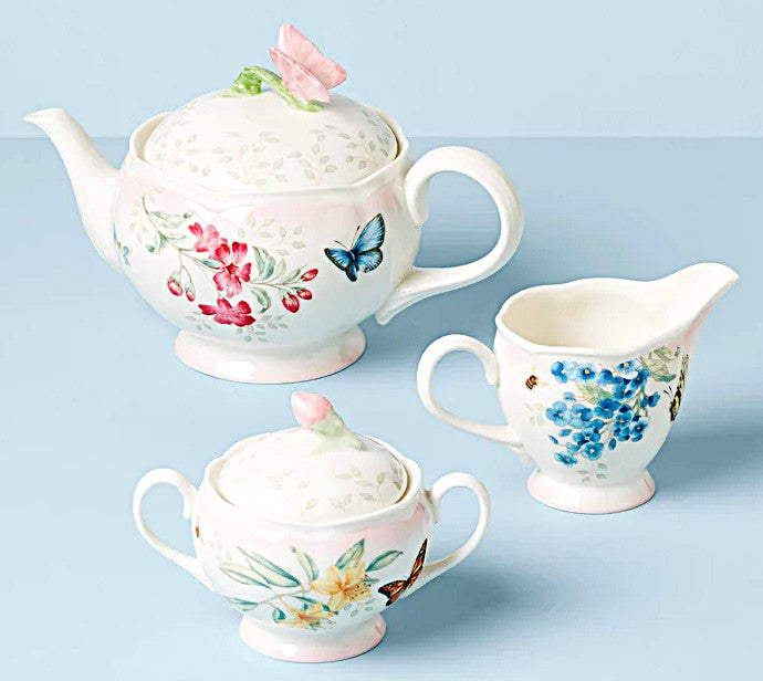 Set of porcelain teapot, sugar bowl, and creamer with floral and butterfly designs on a light blue background.