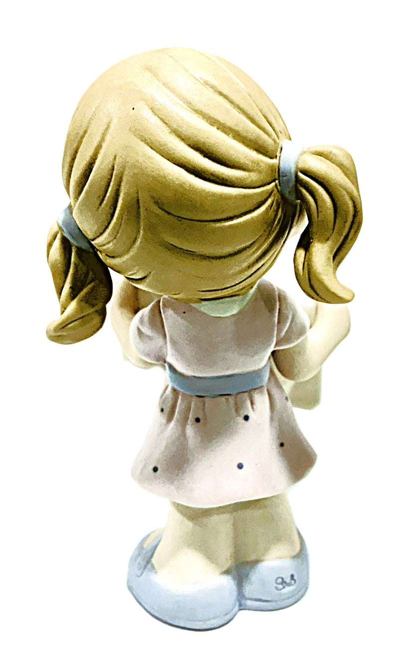 A ceramic figurine of a young girl with blonde pigtails, viewed from behind. She is wearing a light pink dress with small dark polka dots and a light blue waistband and shoes.