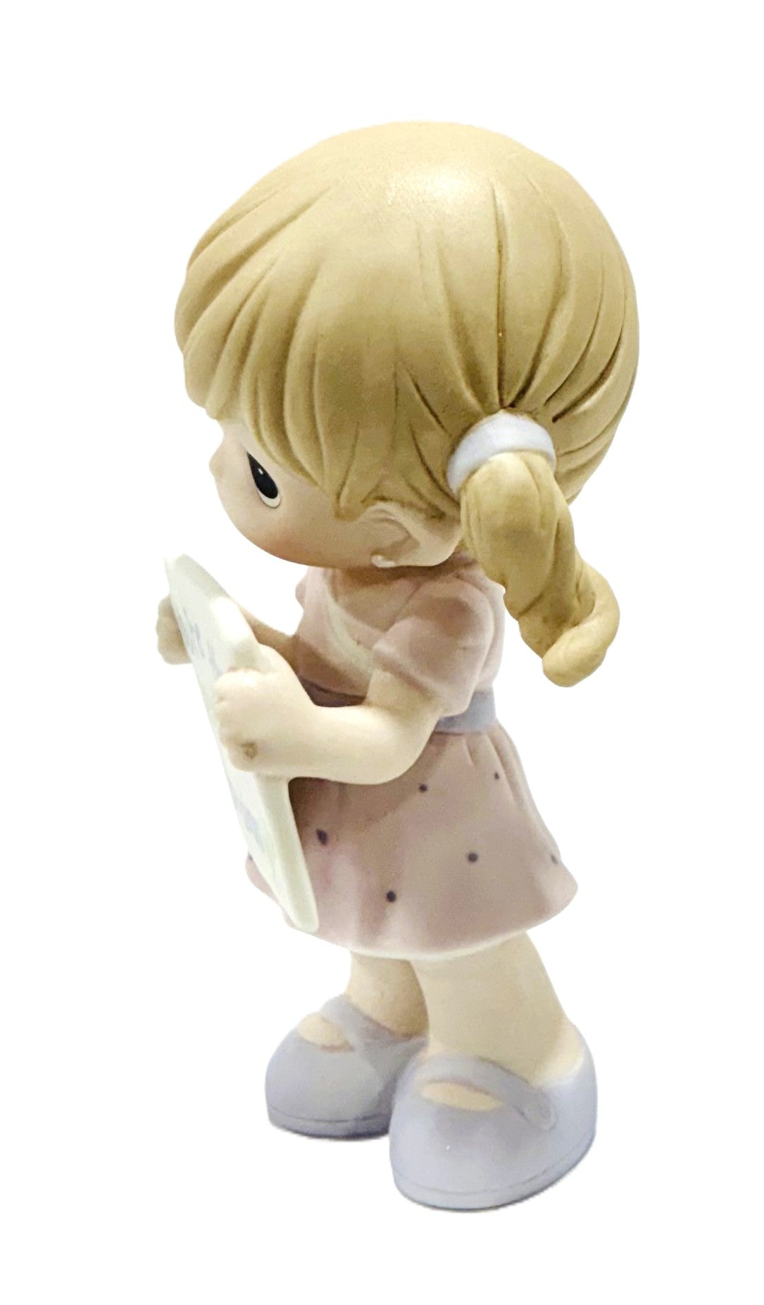 A small figurine of a blonde girl with pigtails, wearing a light pink dress with tiny dark polka dots and holding a white object, shown against a plain white background.